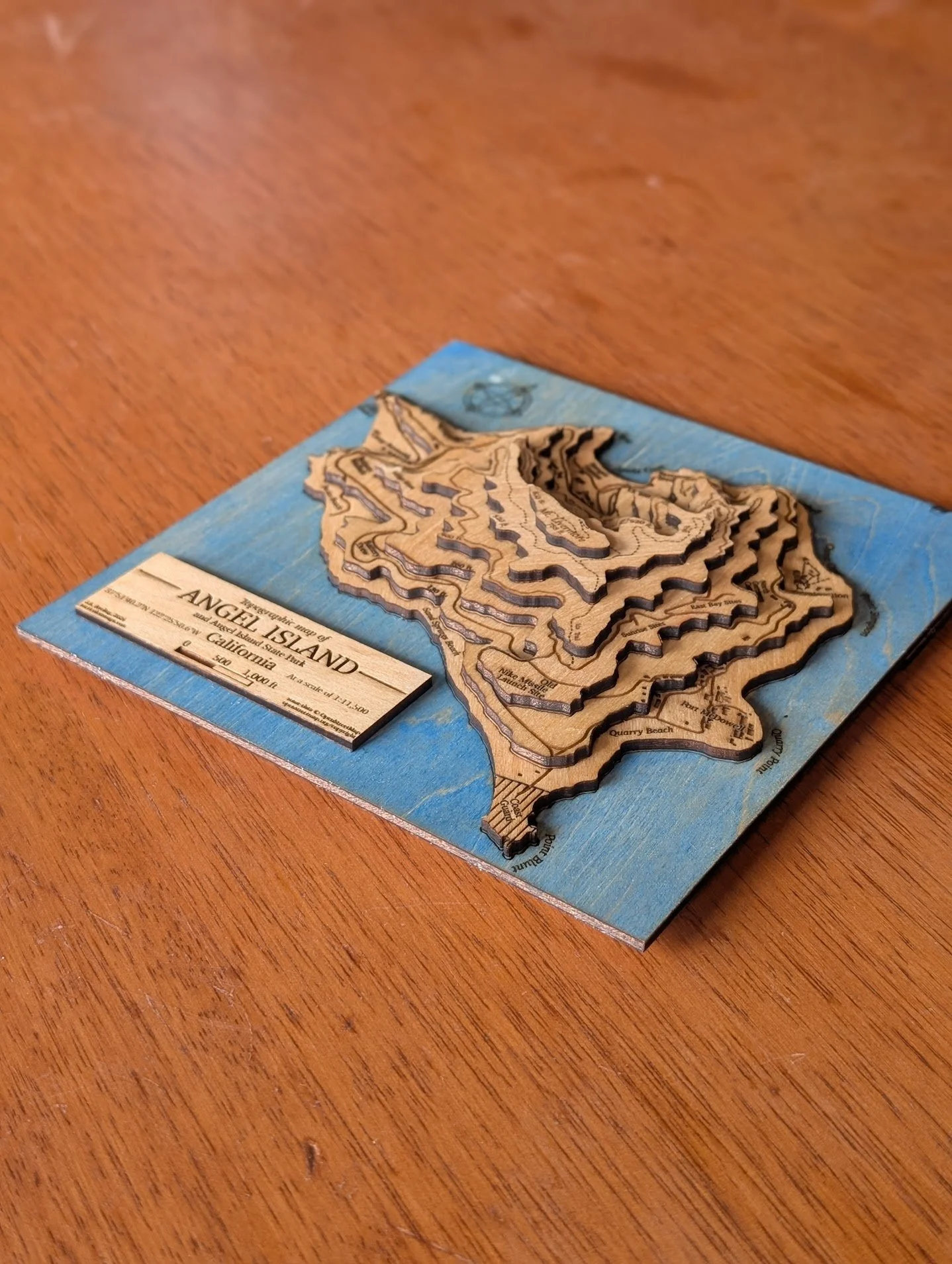 The Angel Island map viewed from a low angle, showing the depth of the stacked wood layers and the three-dimensional terrain. A separate title plaque reading 'Angel Island, California' with scale information sits beside the map.