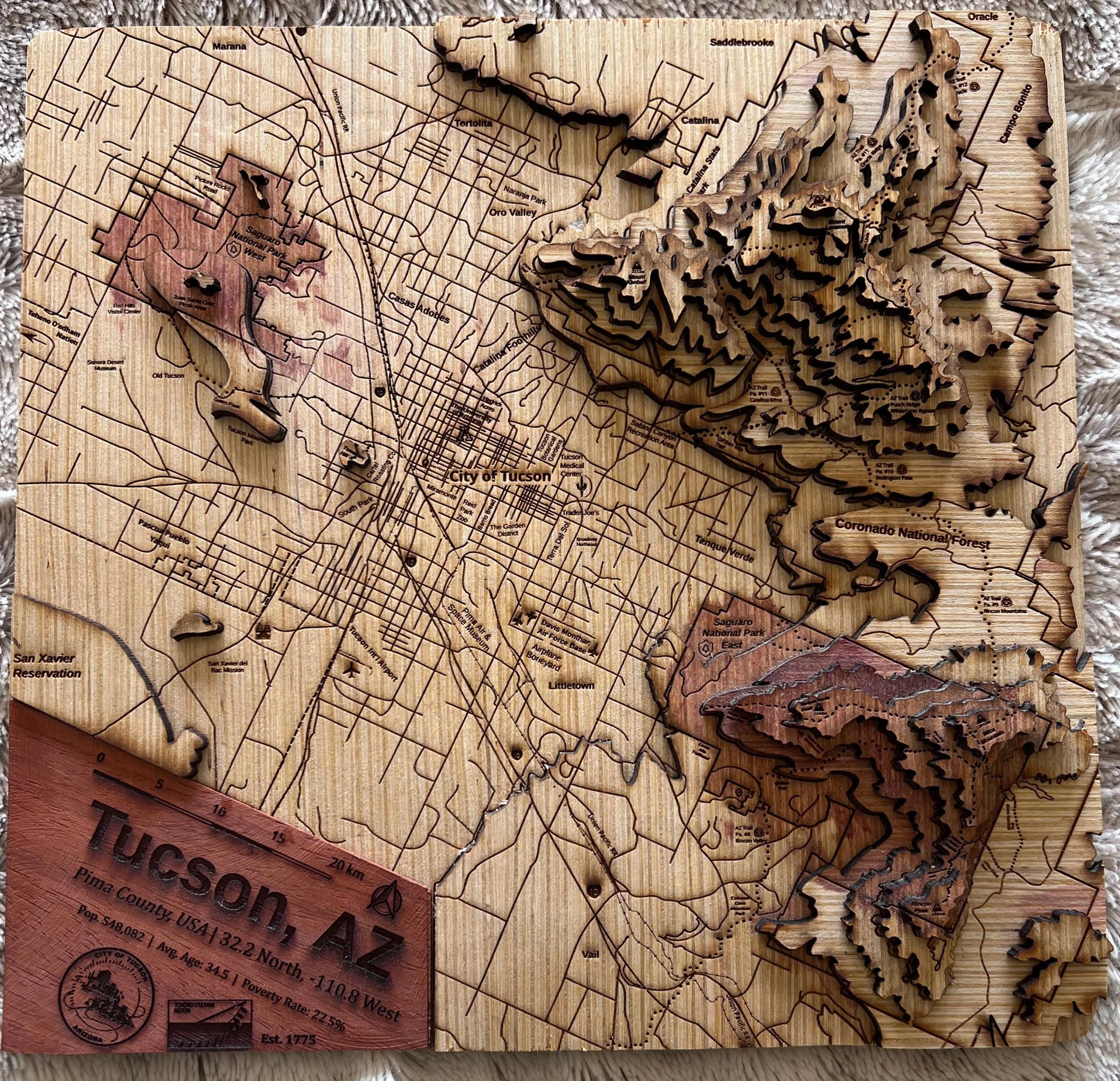 The same Tucson map from a different angle, showing the full composition with the engraved street grid, the deeply stacked mountain layers, and the reddish padauk inlays marking the national park boundaries.